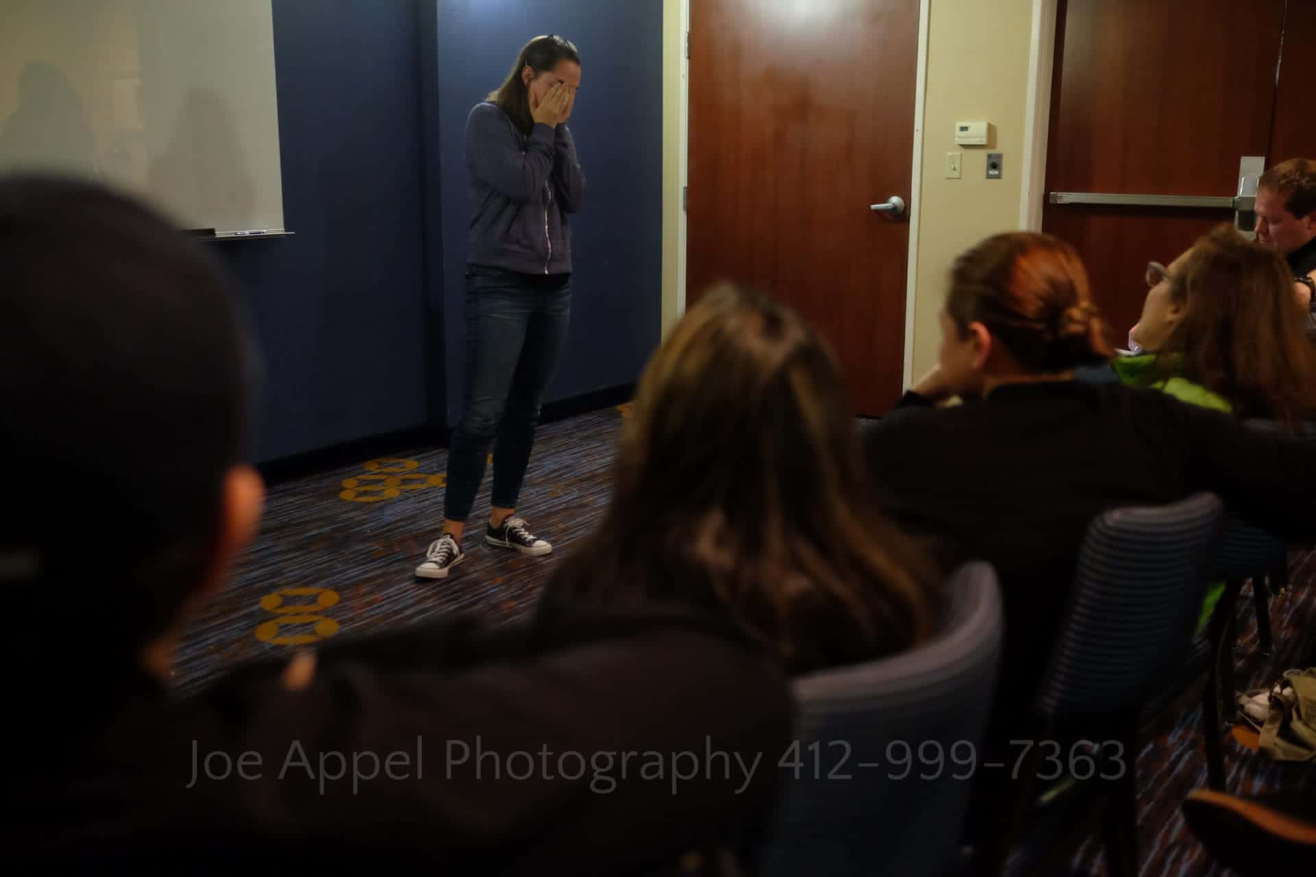 a woman holds her hands to her face as she stands in front of a group of people seated in a conference room.