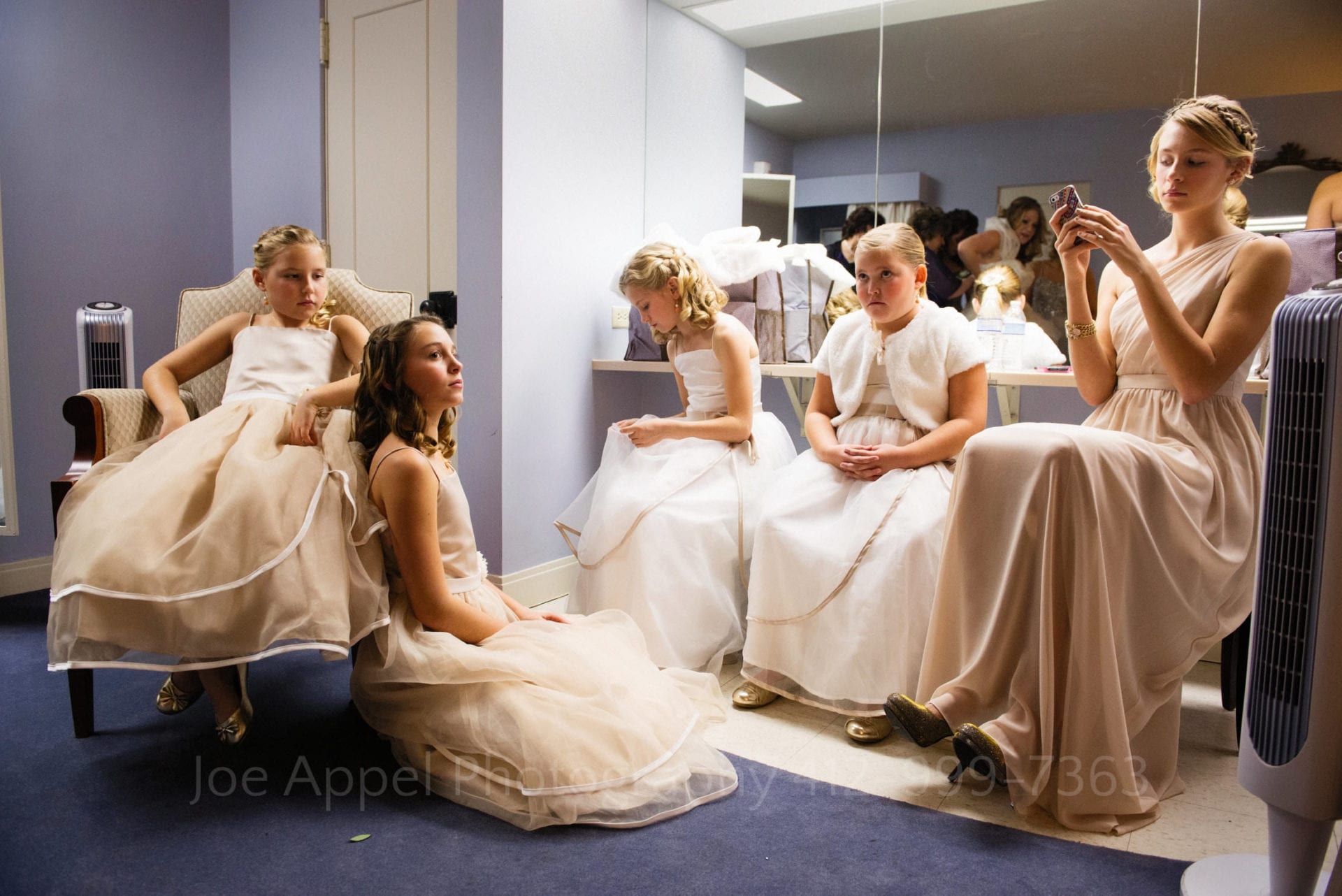 A group of young girls wearing long dresses sit in a lavender waiting room with a mirror behind them.