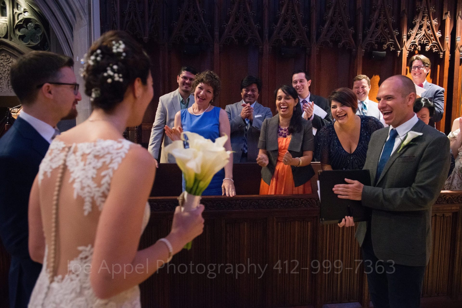 A bride and a groom walk towards a group of people in a choir seated in pews. The choir members smile at the couple who are also happy to see them.