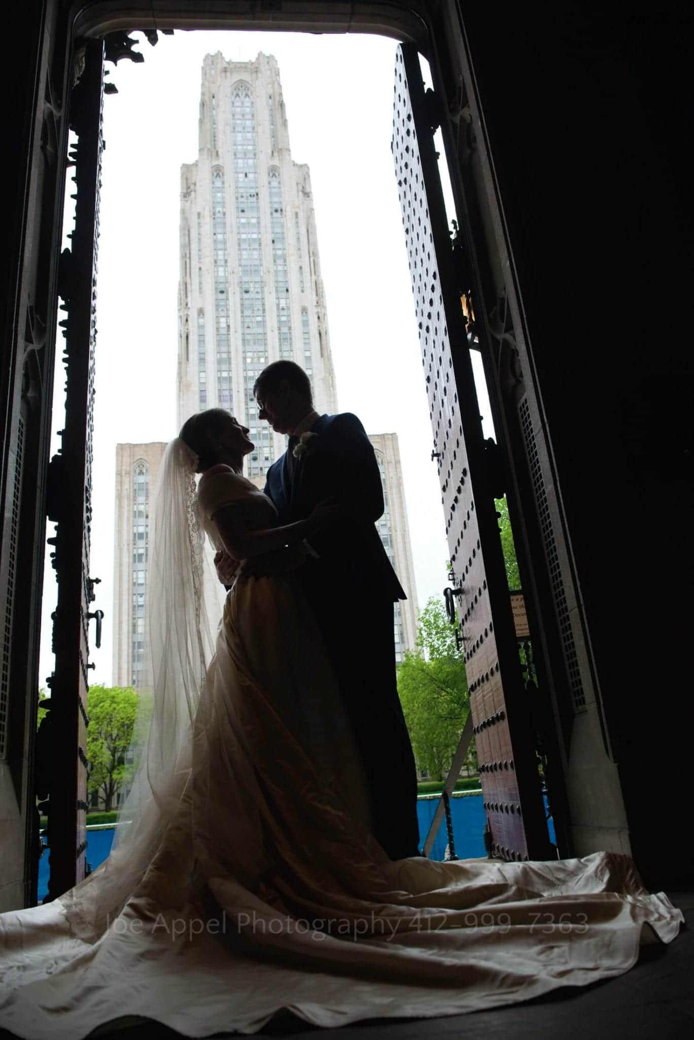 silhouetted in the door at Heinz Chapel weddings, a couple faces each other. The Cathedral of Learning is in the background. 