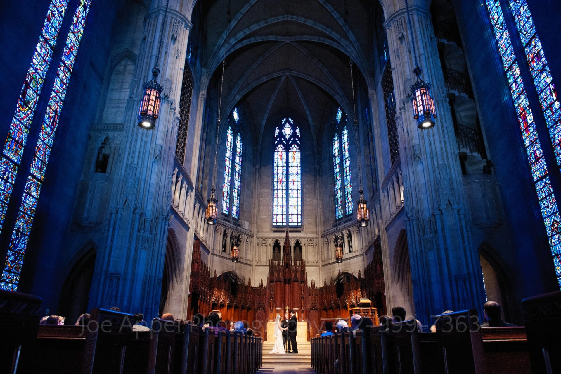 expansive shot of the interior of heinz chapel. The stained glass windows let in purple and blue light while the couple is in warm light in the center.