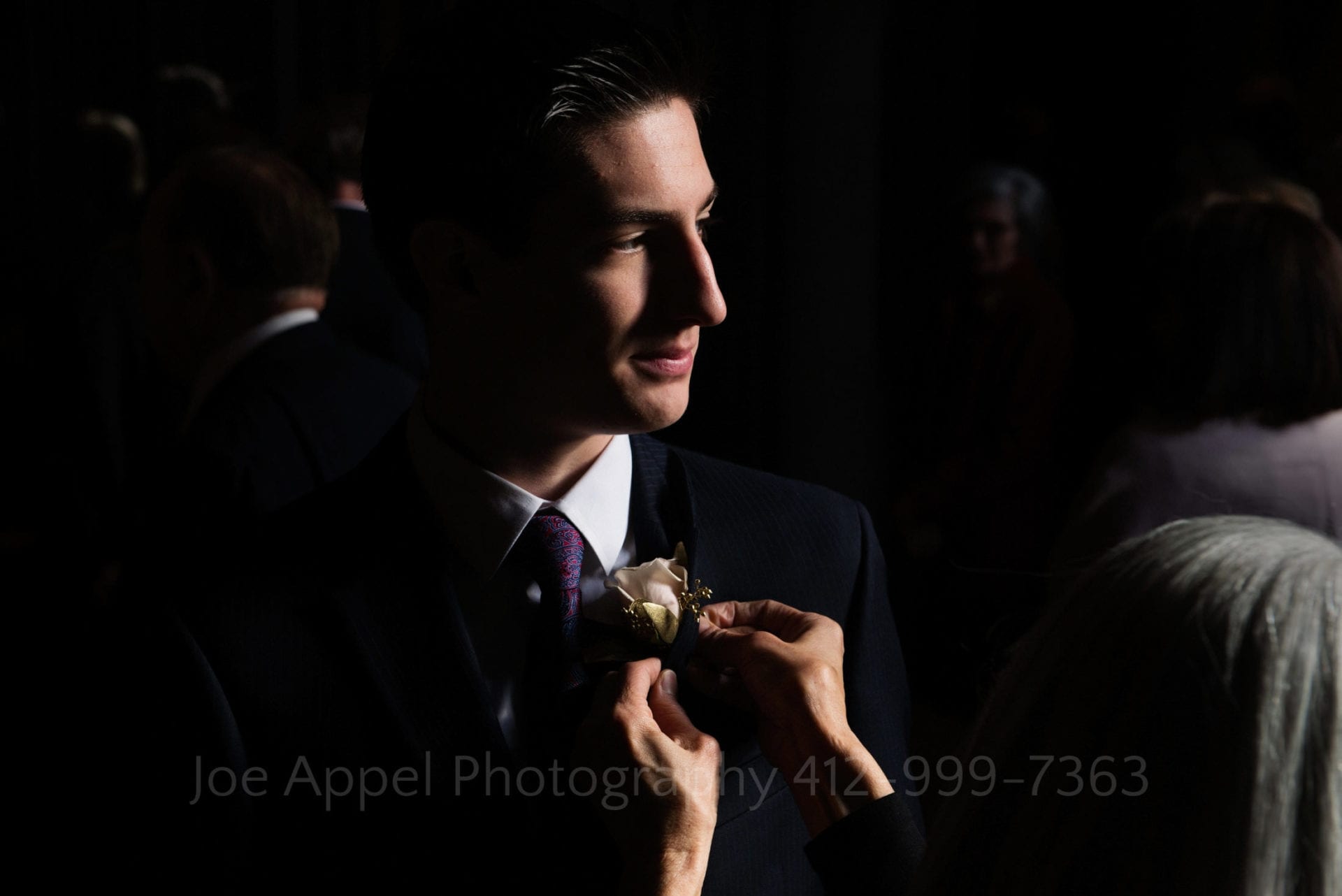 a dramatically lit man in a suit has a boutonniere pinned on his lapel. 