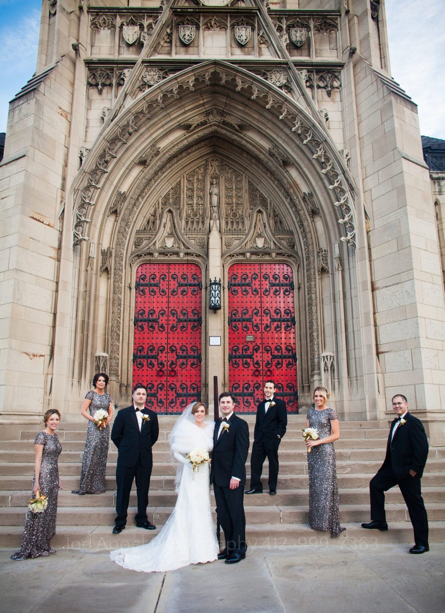 A bridal party stands on the steps at the back of heinz chapel. carved stone and red doors in the background