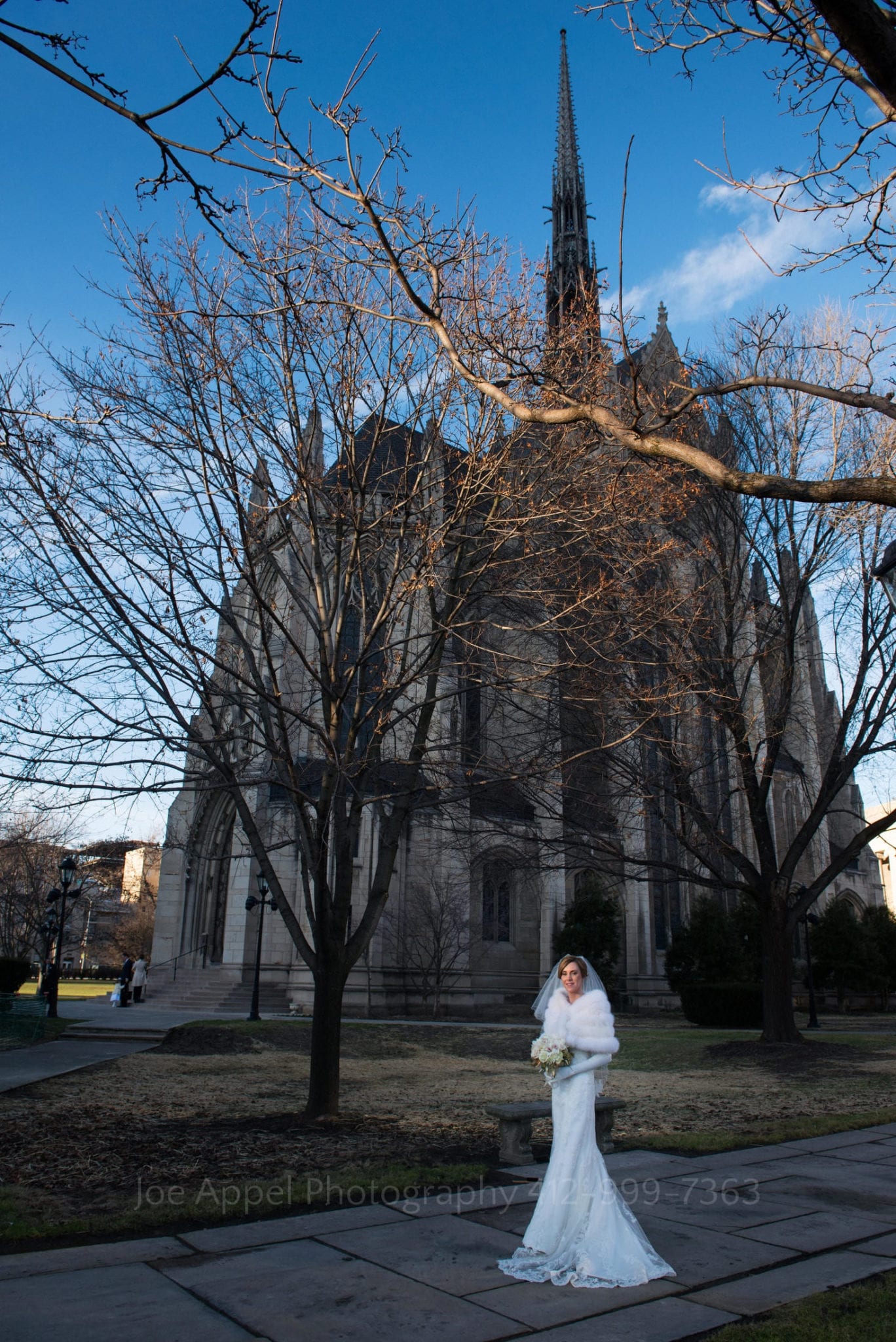a bride wearing a fur wrap and white gloves stands in a shaft of sunlight with heinz chapel visible through the leafless trees behind her.