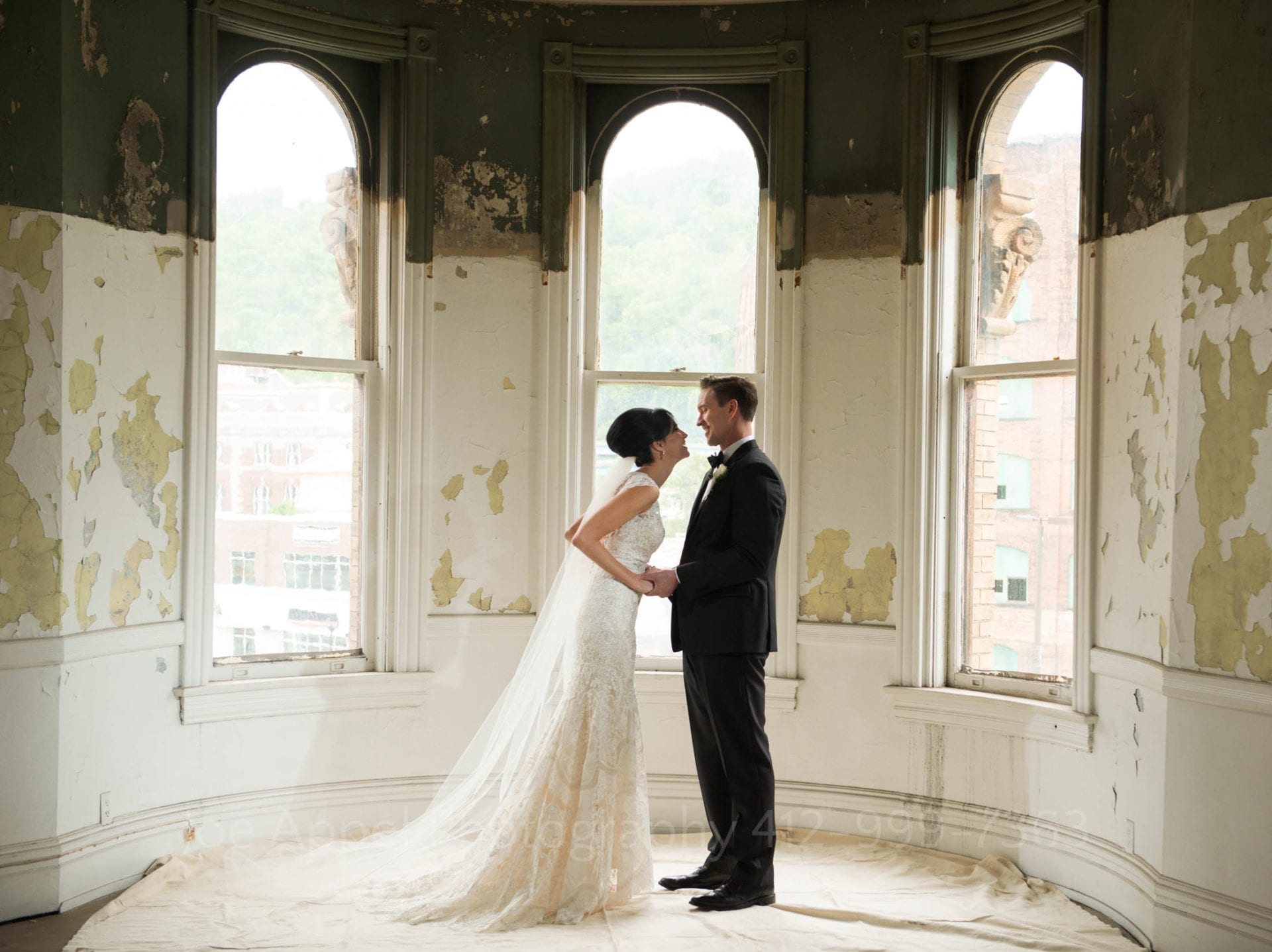 Three arched windows in the background of a bride and groom facing each other in a room with chipped paint. Wedding Photography In West Virginia.