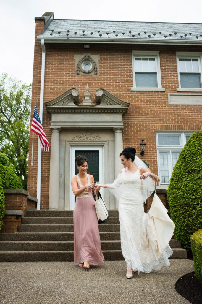 A bride holds her dress train up as she walks down the steps of a brick house with a woman in a pink gown.