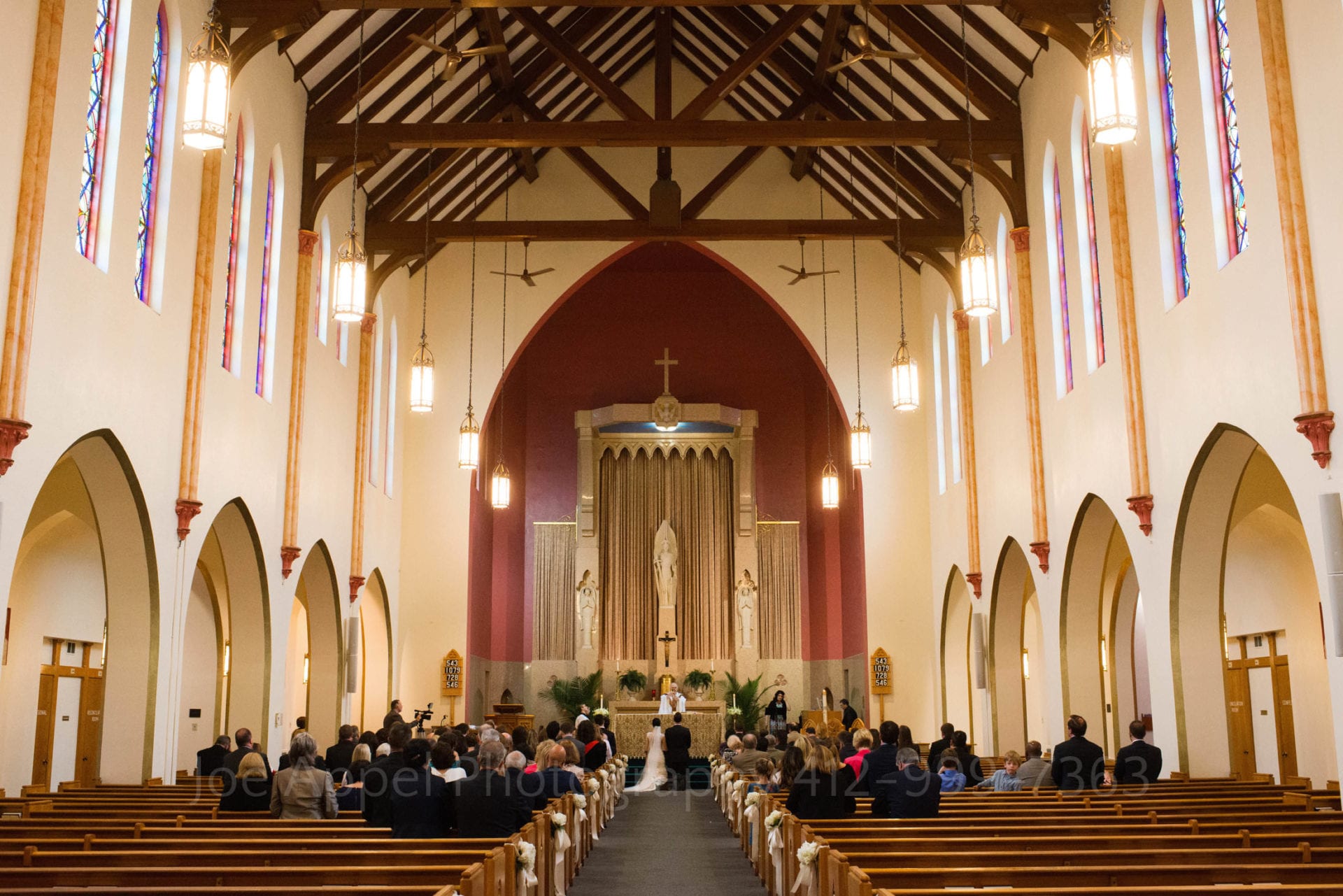 the interior of a catholic church during a wedding. the walls of the church are white while the area behind the altar is red. There are wooden beams exposed on the ceiling.