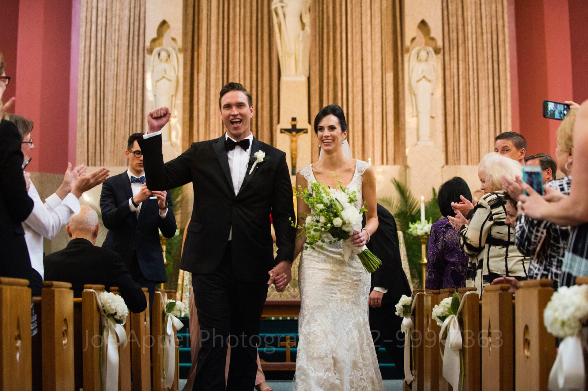 a groom raises his fist in the air as he and his bride start to make their way down the aisle of a church at the end of their wedding ceremony