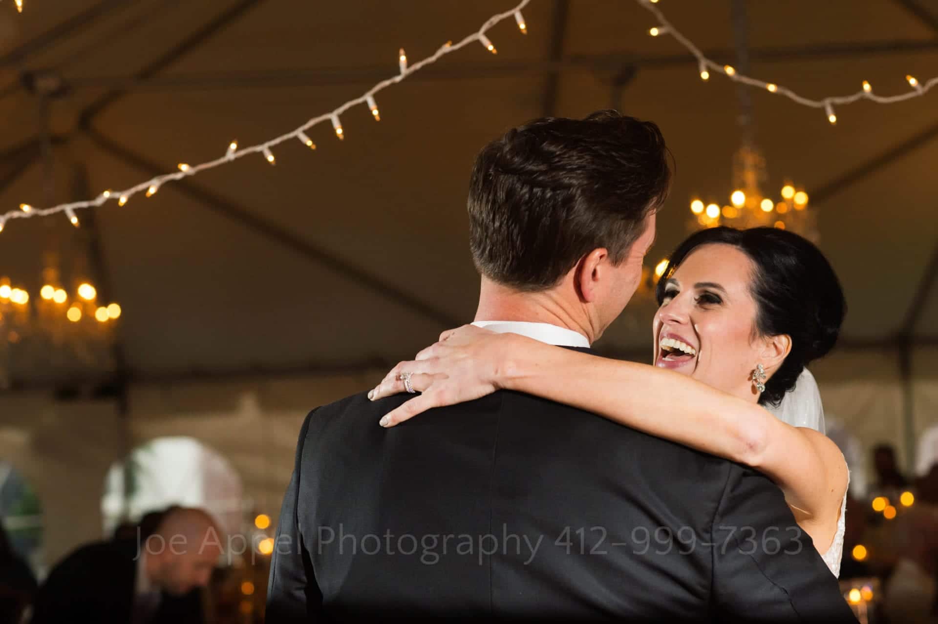 A bride smiles broadly at her groom as they dance. Her arm is around his neck.