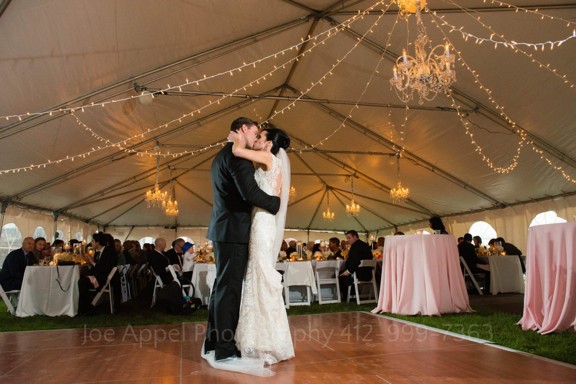 wide shot of a bride and groom dancing on a wooden floor inside of a decorated tent. Chandeliers and strings of twinkle lights illuminate.