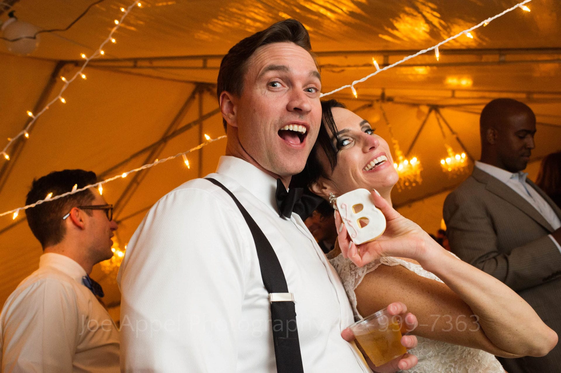 A bride leans into her groom as she holds a cookie in the shape of the letter B. They're smiling at the camera. Wedding Photography In West Virginia.