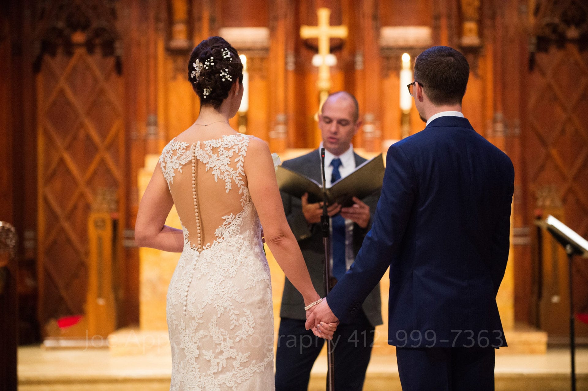 Seen from behind, a bride and groom hold hands on the altar during their Heinz Chapel and LeMont Wedding.