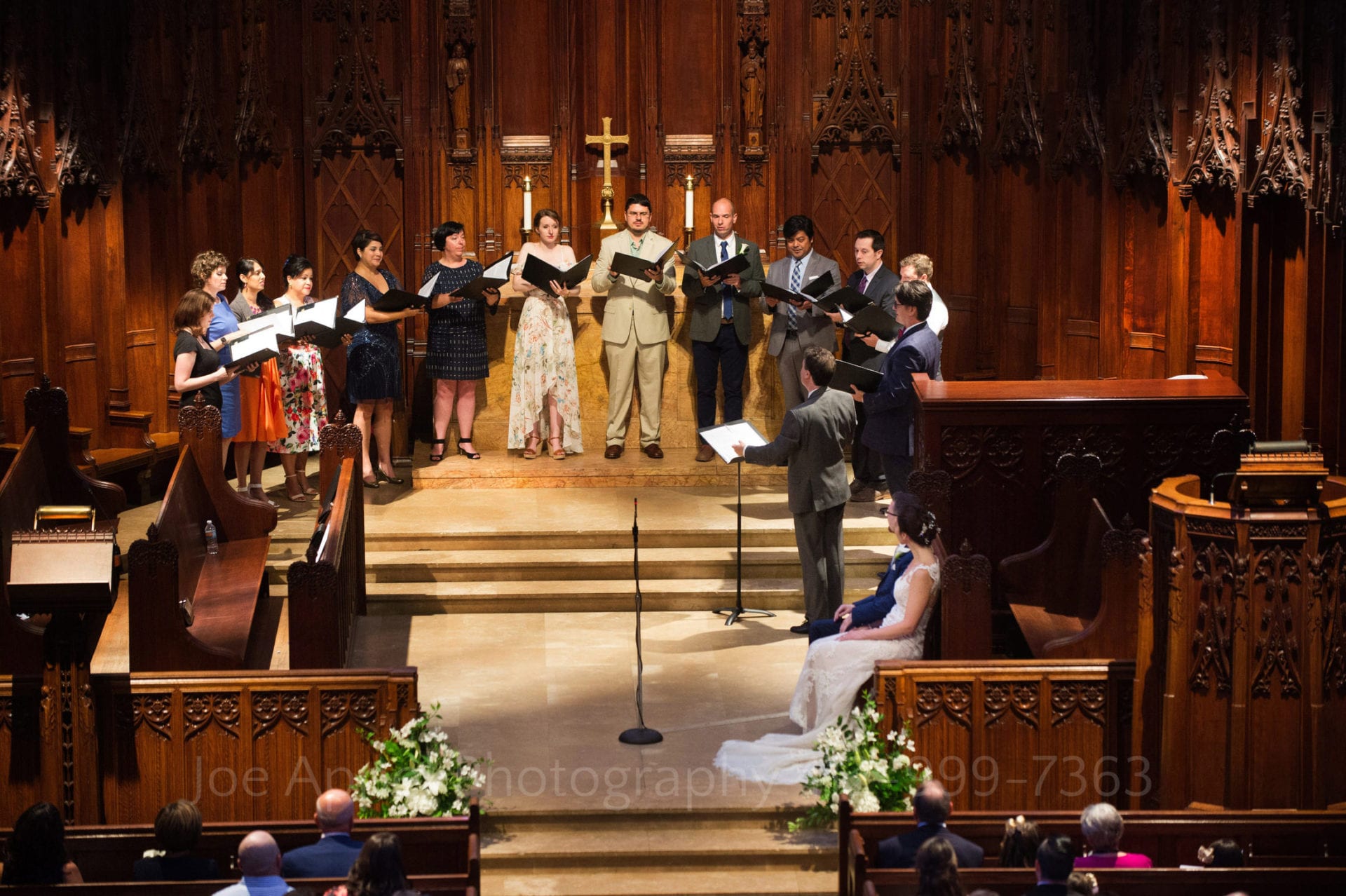 A choir stands in a semi-circle on the altar at Heinz Chapel as the bride and groom sit listening to them.