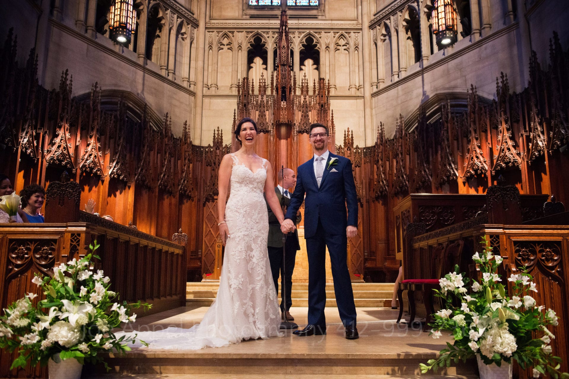 A bride and groom stand holding hands as they are presented to the audience at the end of their Heinz Chapel and LeMont Wedding.