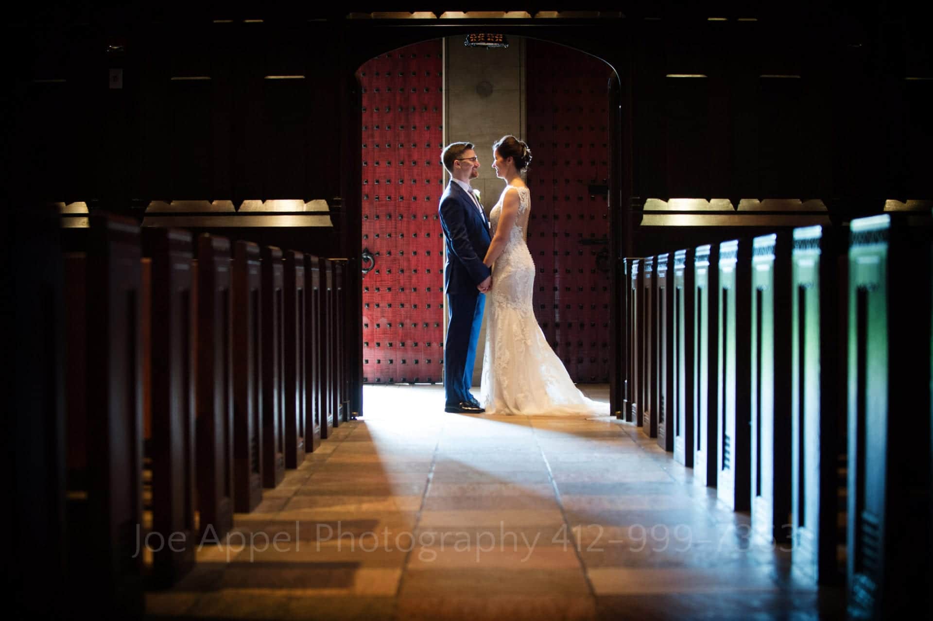 Standing at the back of the church, a bride and groom are rim lit from the open door behind them.