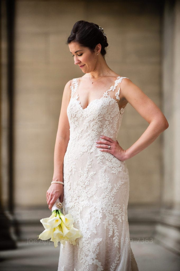 In a stone portico, a bride stands looking down at her bouquet in her right hand. Her left hand is on her hip.
