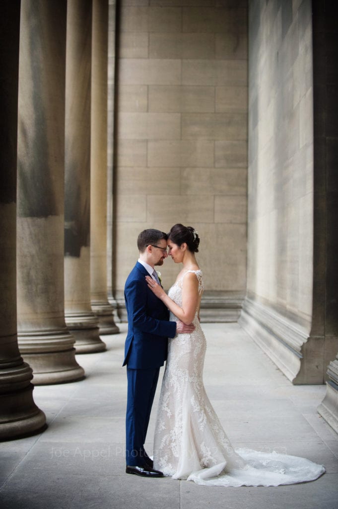 A bride and groom stand in a portico with stone columns. They're facing each other with their foreheads touching during their Heinz Chapel and LeMont Wedding.