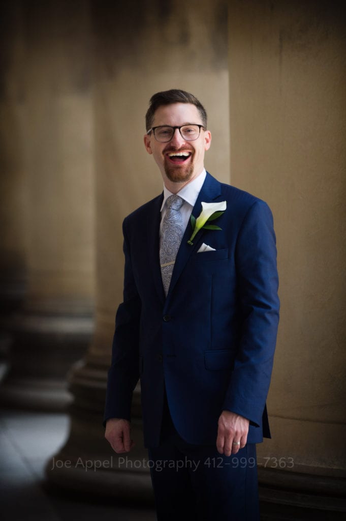 A bearded groom in a blue suit wearing glasses laughs with stone columns behind him during his Heinz Chapel and LeMont Wedding