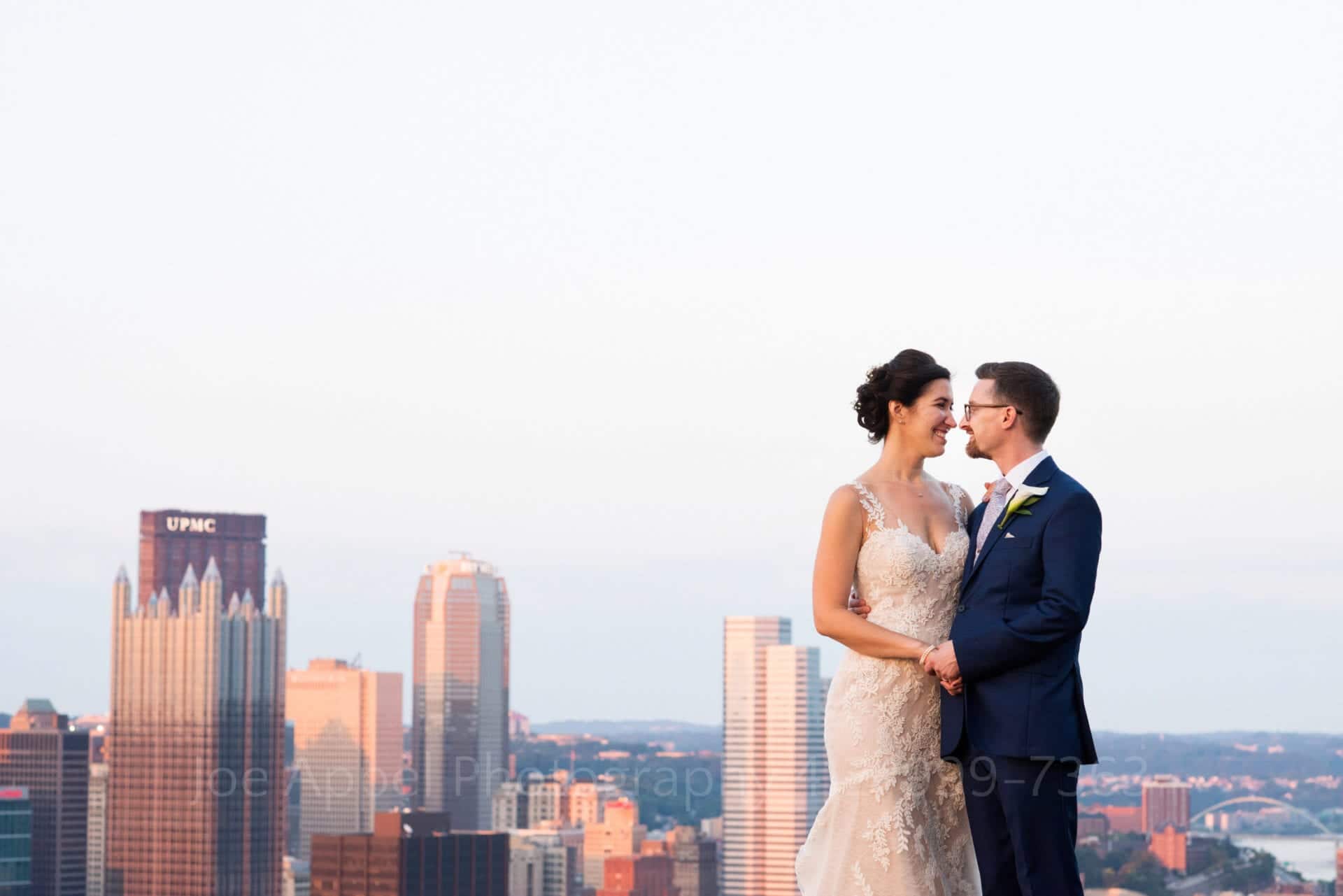 A bride and groom face each other as they stand high above the skyline of the city of Pittsburgh at sunset.