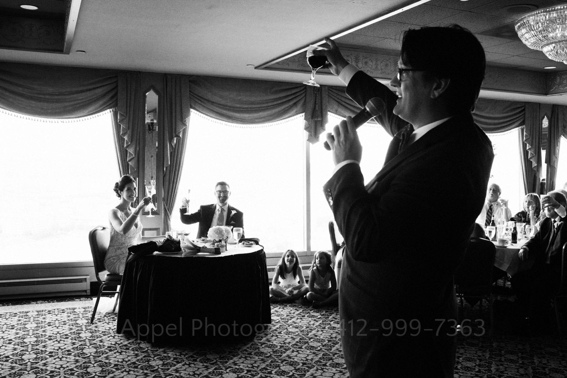 A man lifts a glass of wine as he toasts a bride and groom seated at a table in front of large windows.