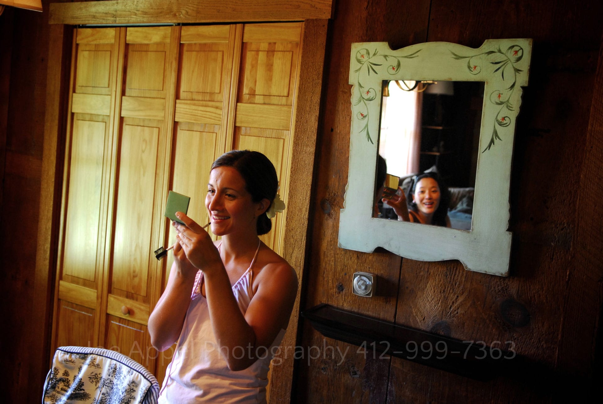 a bride applies lipstick while looking at herself in a compact. Another woman is reflected in the mirror behind her. Armstrong Farms Weddings