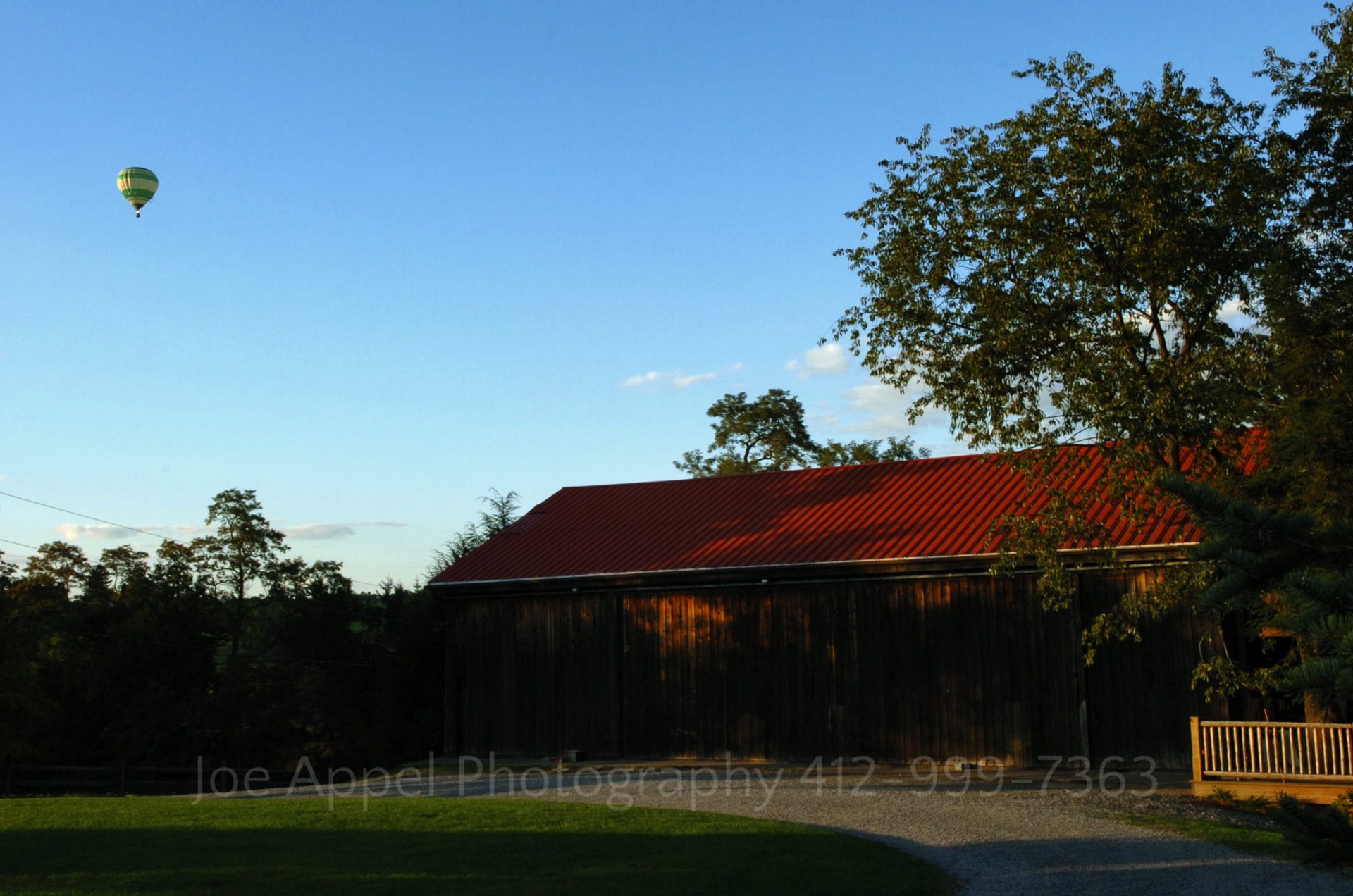A hot air balloon floats in the blue sky high above a red-roofed barn. Armstrong Farms Weddings