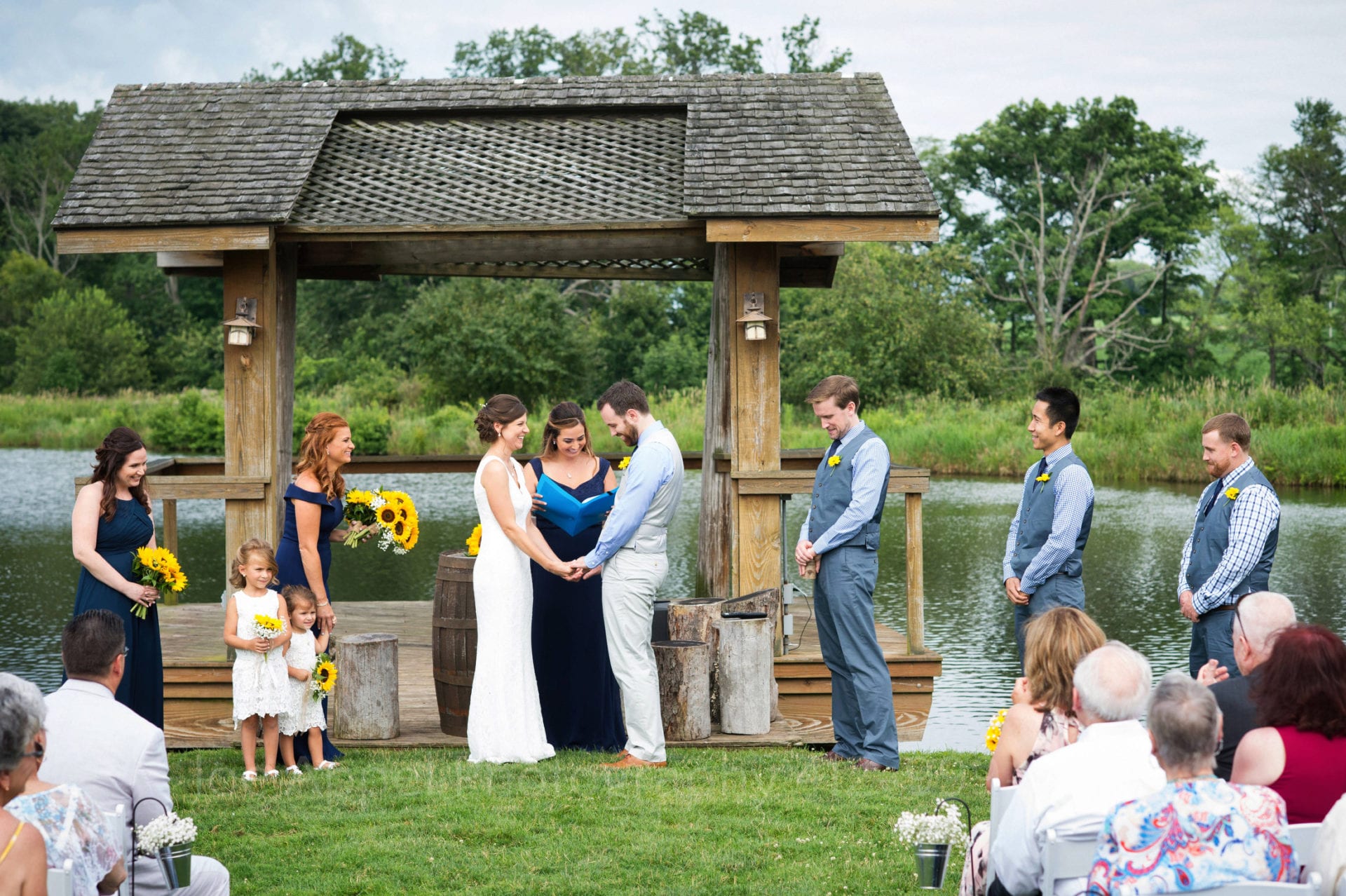 A bride and groom laugh while holding hands as they exchange vows. The backdrop is a pavilion on the edge of a pond. The bridal party stands on either side of the bride and groom. Armstrong Farms Weddings