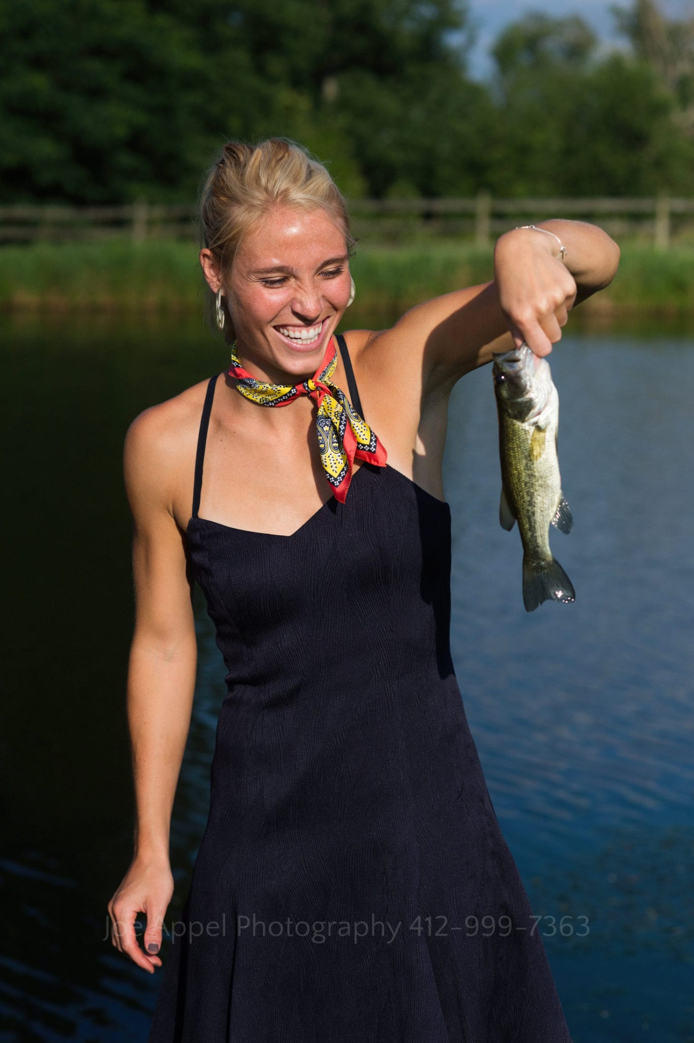 A smiling blond woman wearing a dark blue dress holds a fish that she caught from the pond behind her. Armstrong Farms Weddings