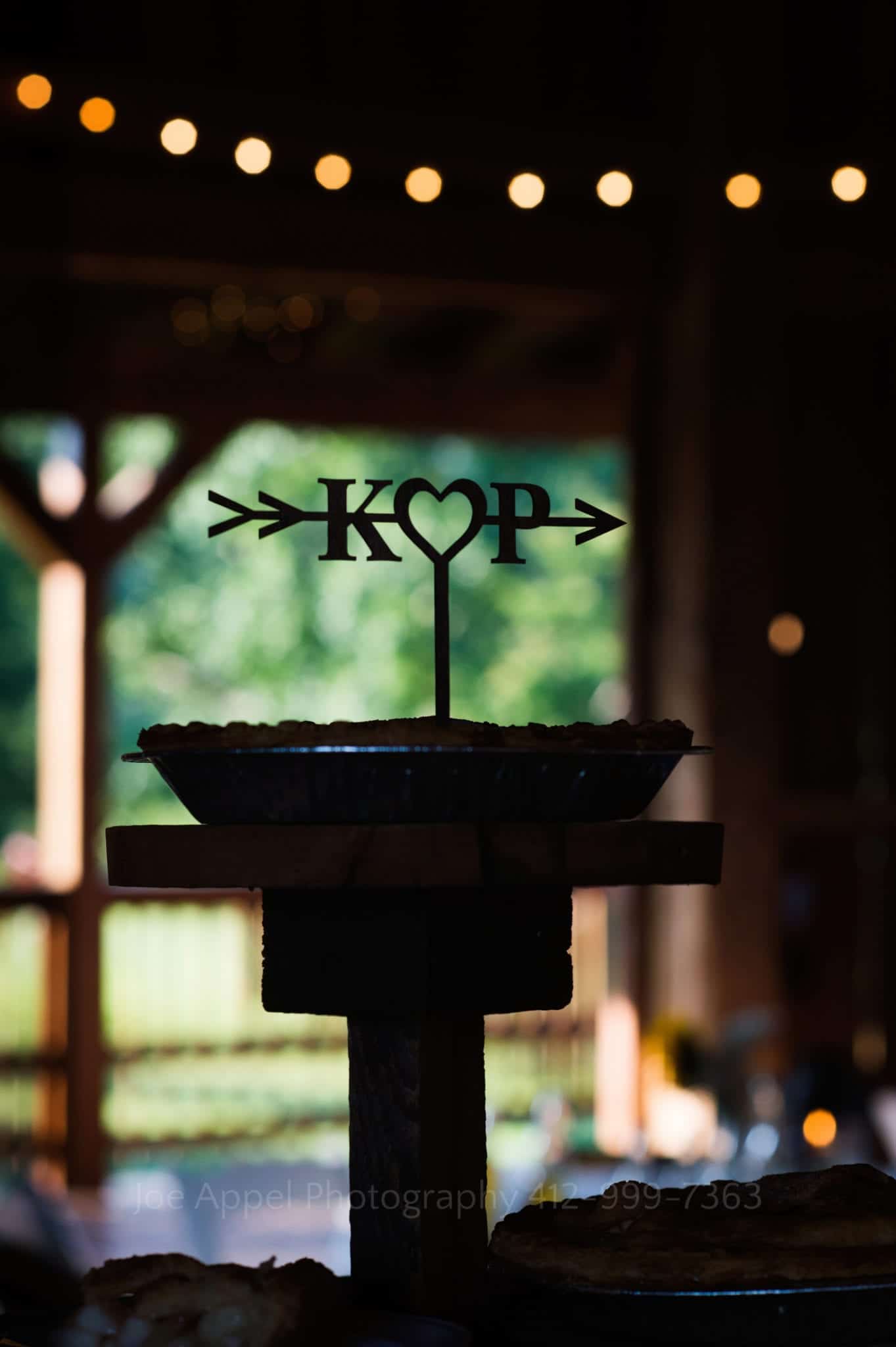 The letters K and P with a heart between them and an arrow through all of it is a topper on a pie seen in silhouette. Armstrong Farms Weddings