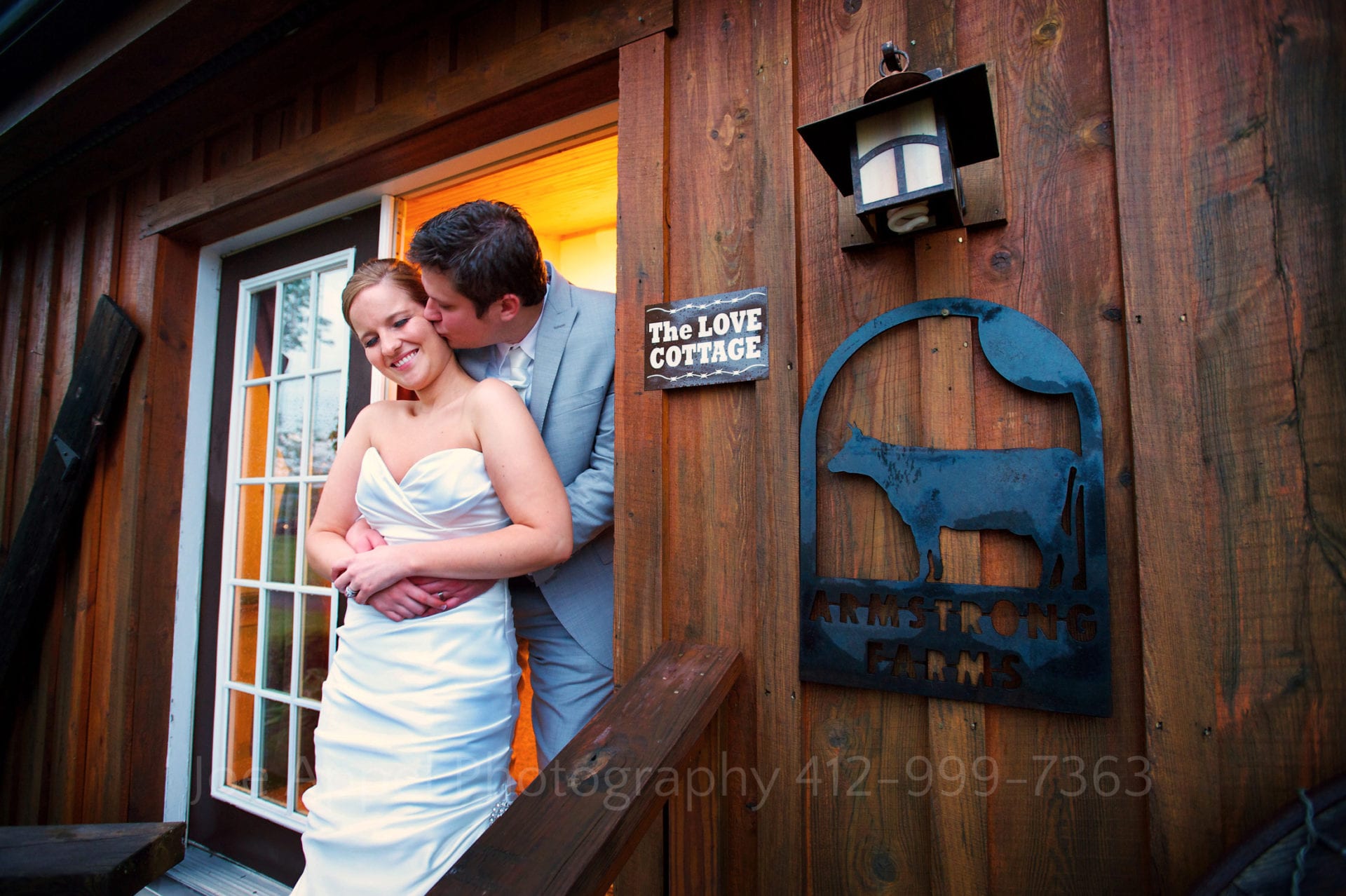 A bride smiles as her groom embraces her from behind and kisses her on the cheek in the doorway of a house called "The Love Cottage" Armstrong Farms Weddings