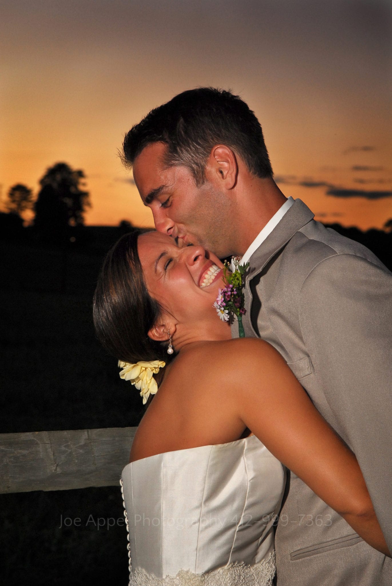 With deep orange sunset behind them a groom hugs his bride tightly and kisses her on the forehead. The bride grins. Armstrong Farms Weddings