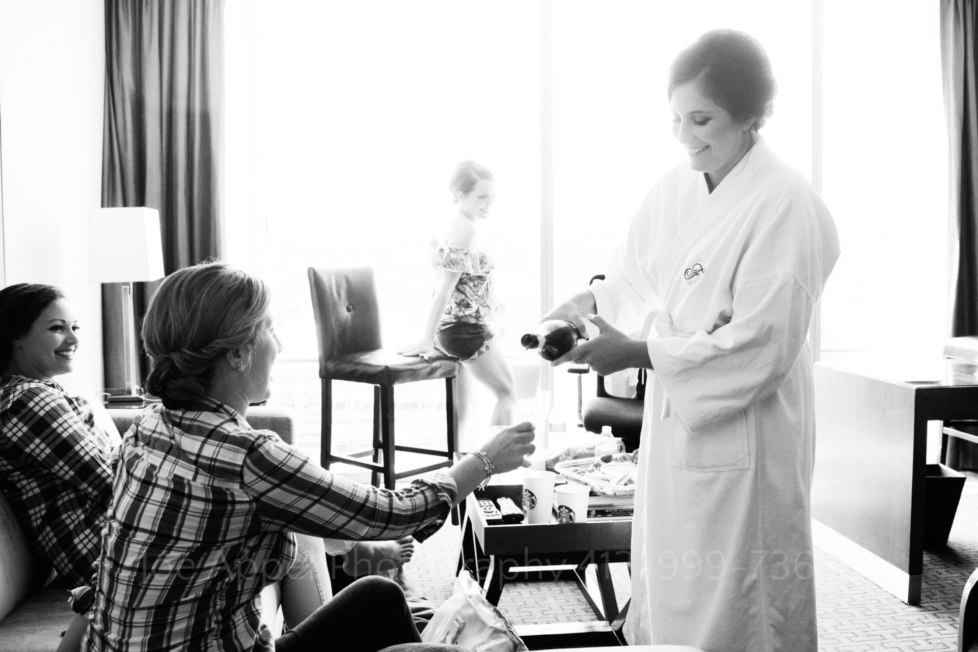A woman wearing a Fairmont bathrobe pours champagne for two other women. A bright floor-to-ceiling window is behind them Fairmont Hotel Pittsburgh Weddings.