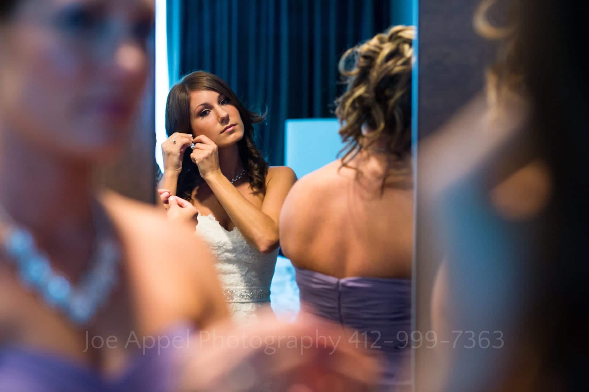 A bride puts an earring on as she looks in the mirror while bridesmaids in purple dresses get ready next to her Fairmont Hotel Pittsburgh Weddings.