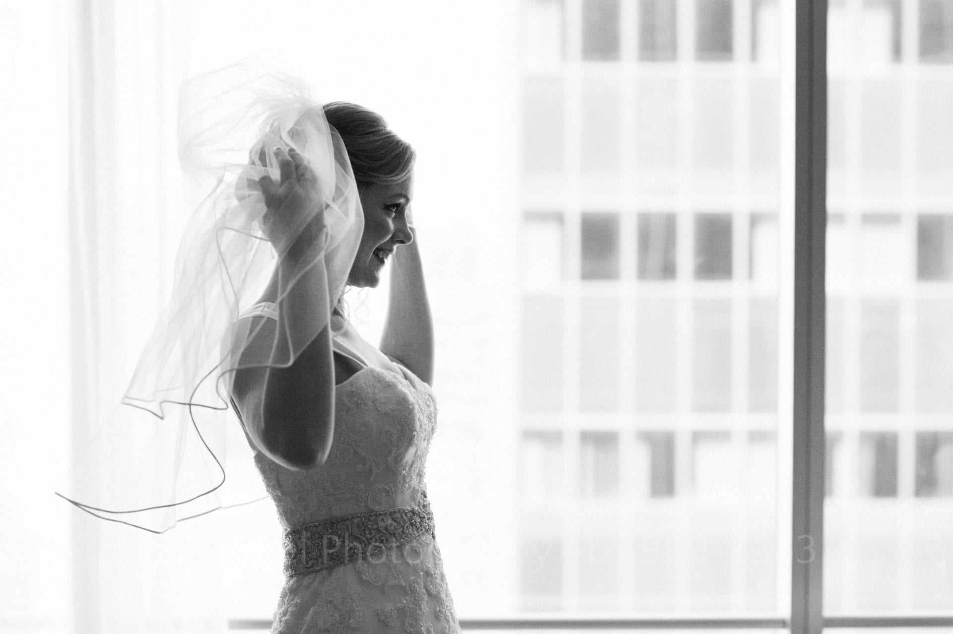 A bride smiles as she fluffs her veil in front of a large window Fairmont Hotel Pittsburgh Weddings.