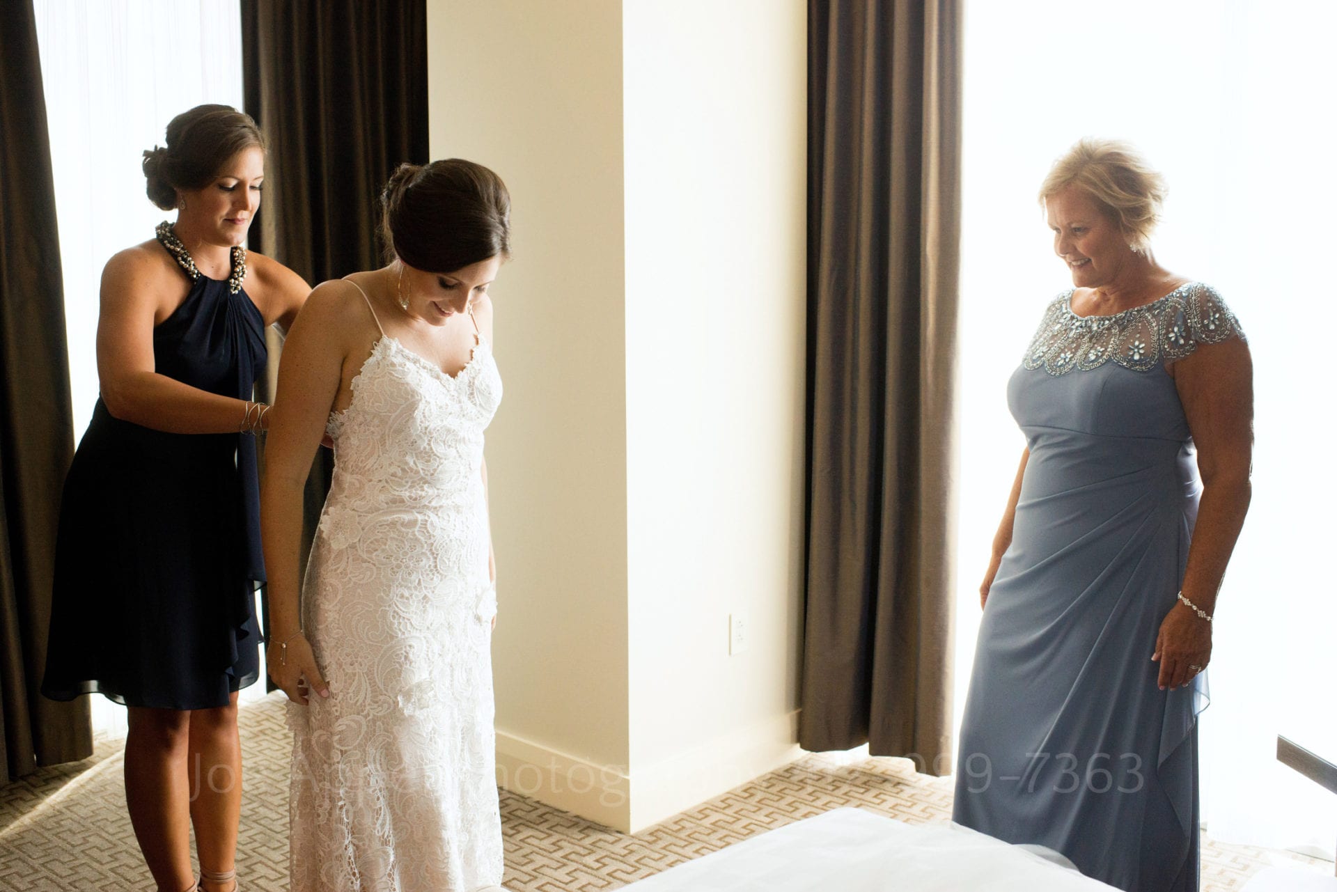 A bride is helped into her dress by a bridesmaid wearing a blue dress as their mother stands watching while wearing a light blue dress Fairmont Hotel Pittsburgh Weddings.