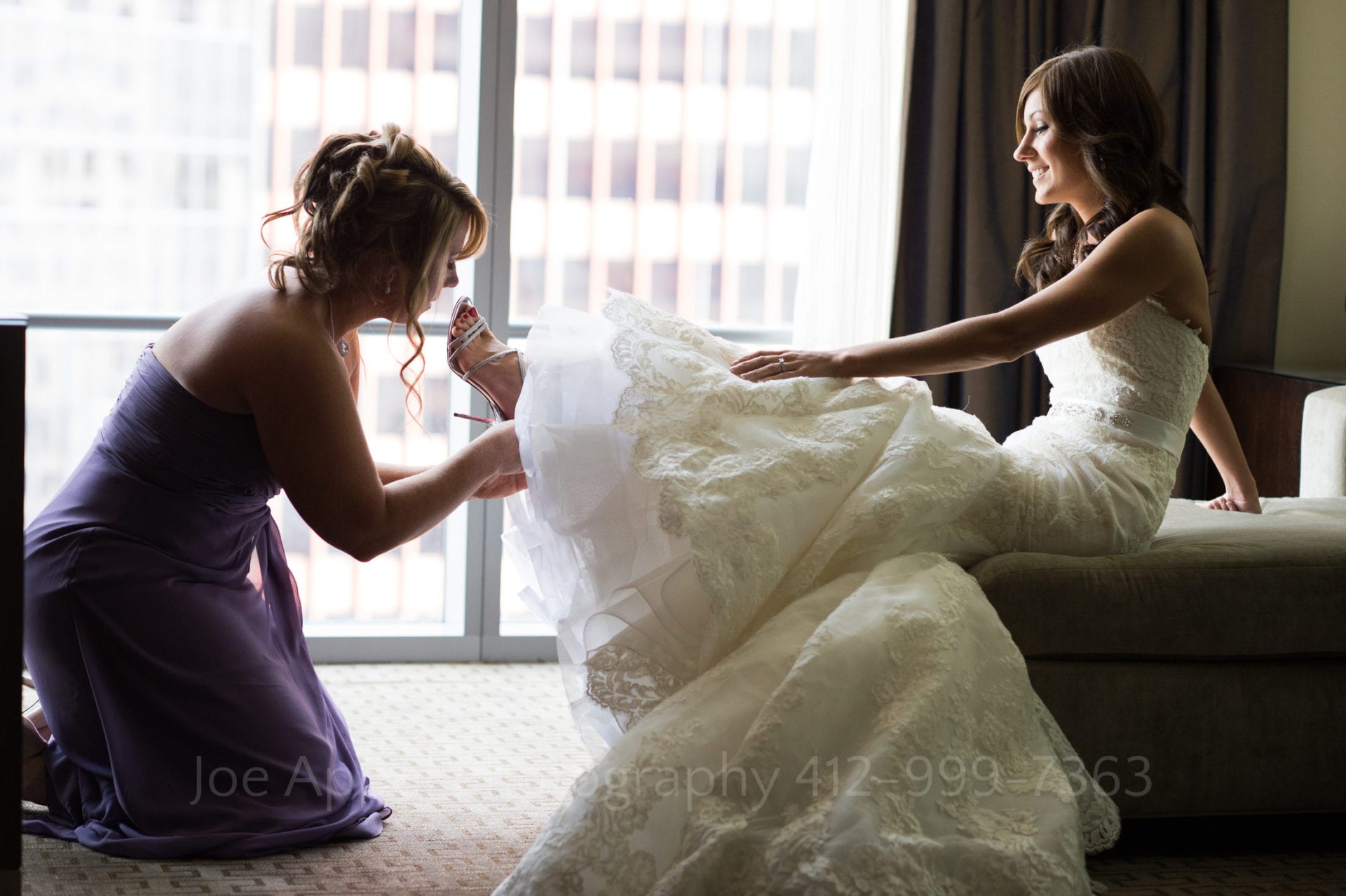 A bride sits on the end of a bed while her bridesmaid helps put on her shoes. The window behind them provides the lighting Fairmont Hotel Pittsburgh Weddings.
