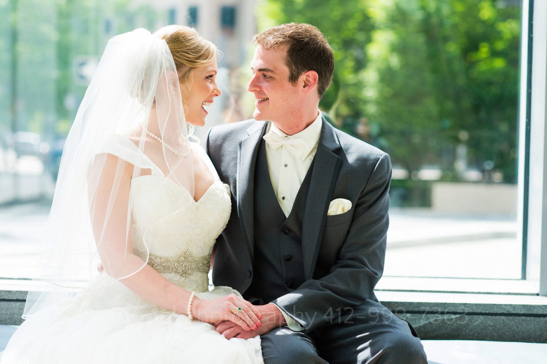 A bride and groom sit together and smile at one another in front of a window on a sunny day Fairmont Hotel Pittsburgh Weddings.
