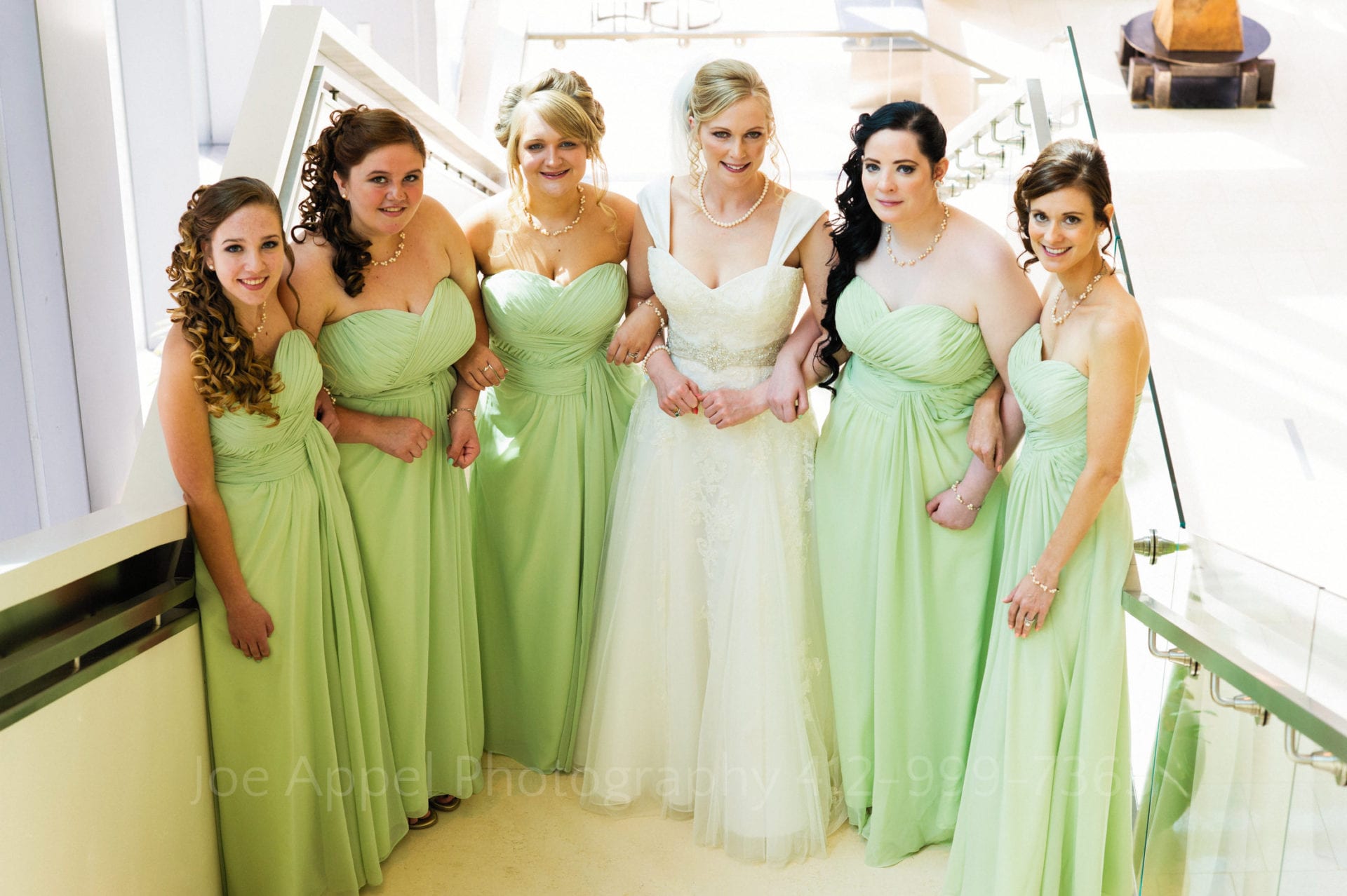 A bride and her green-dressed bridesmaids stand on a lower landing on a stairway Fairmont Hotel Pittsburgh Weddings