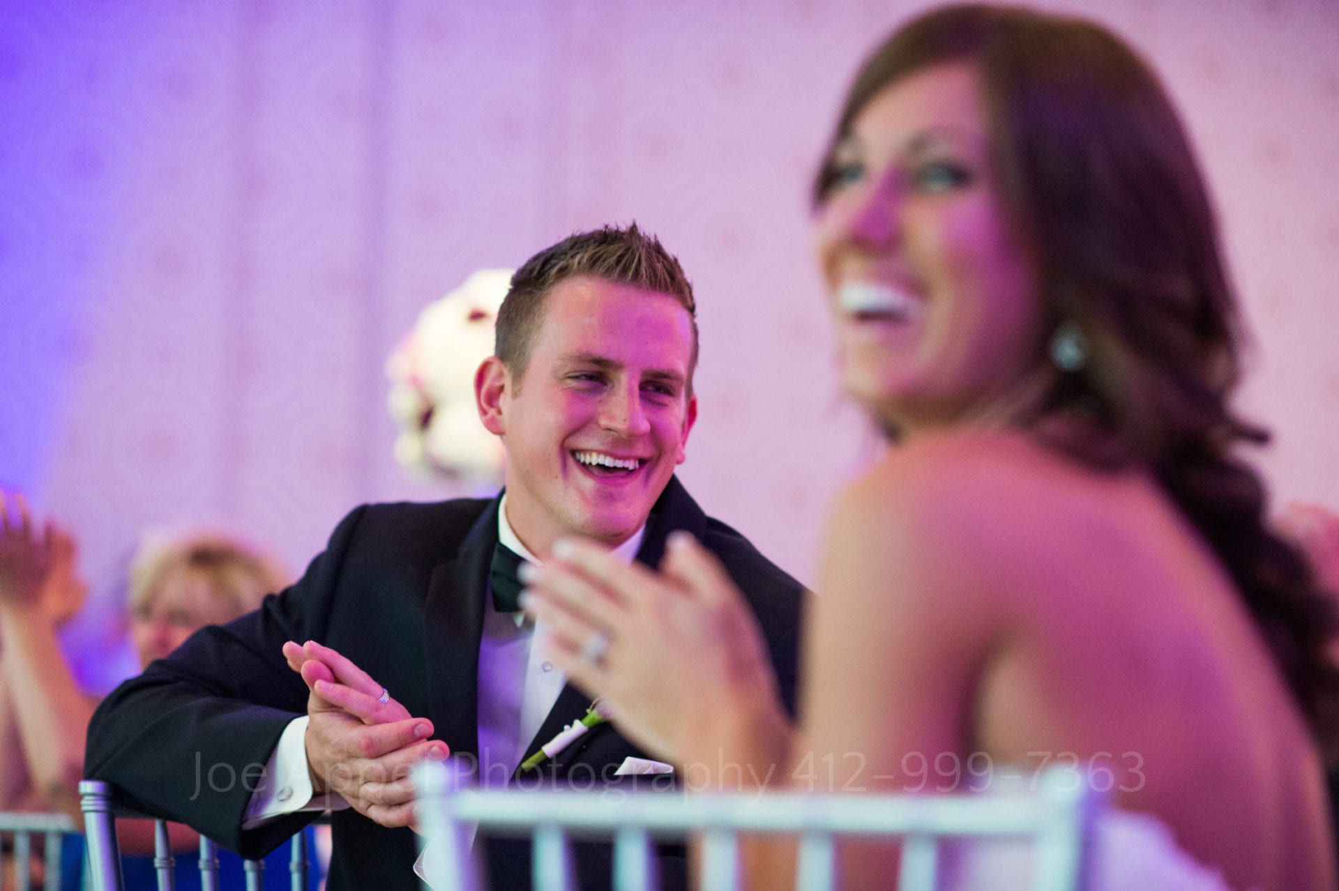 A groom looks at his bride as they both sit in chairs and applaud while listening to speeches Fairmont Hotel Pittsburgh Weddings.