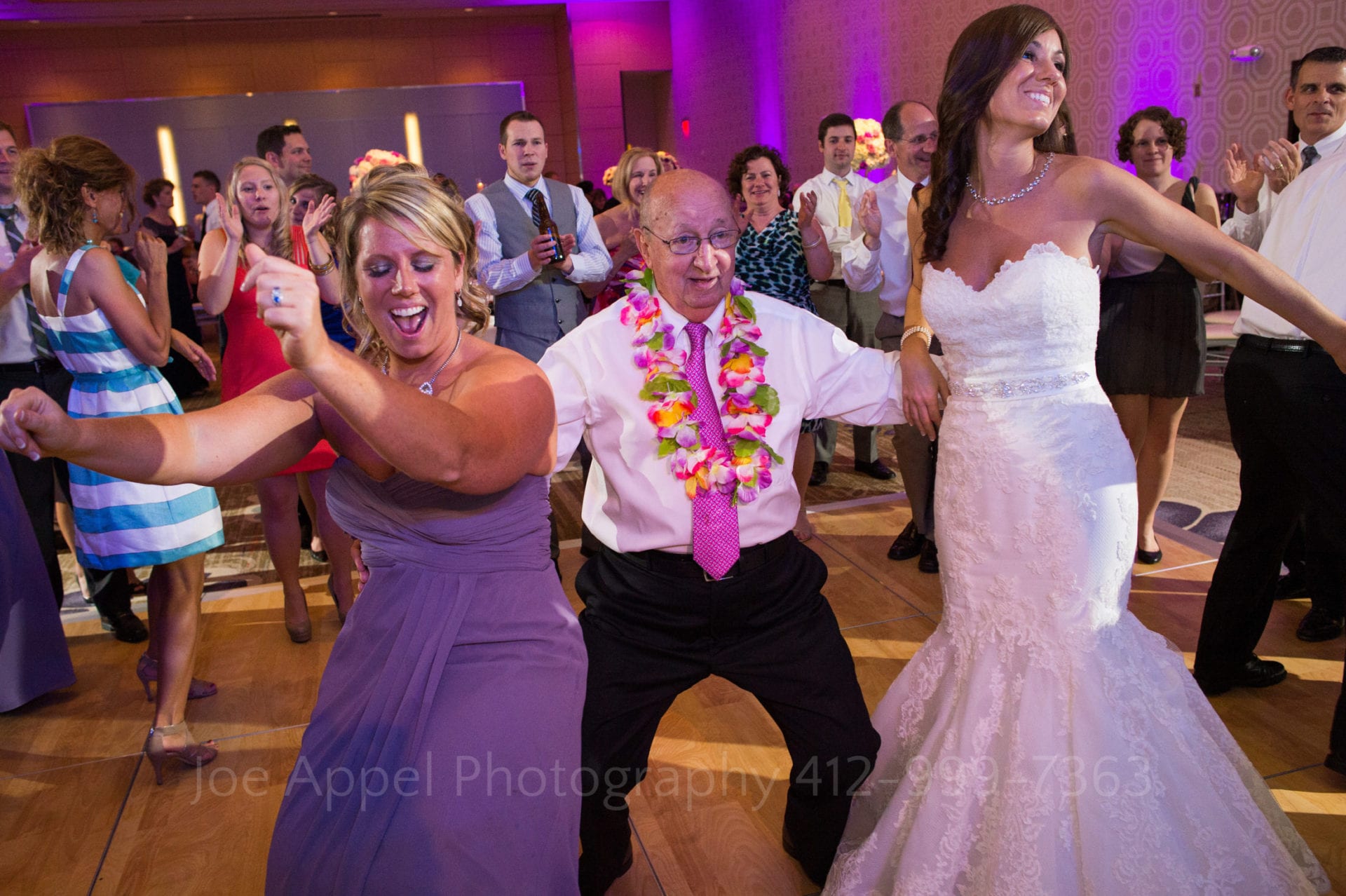 An old man wearing a lei dances with a bride and a bridesmaid in a purple dress Fairmont Hotel Pittsburgh Weddings.