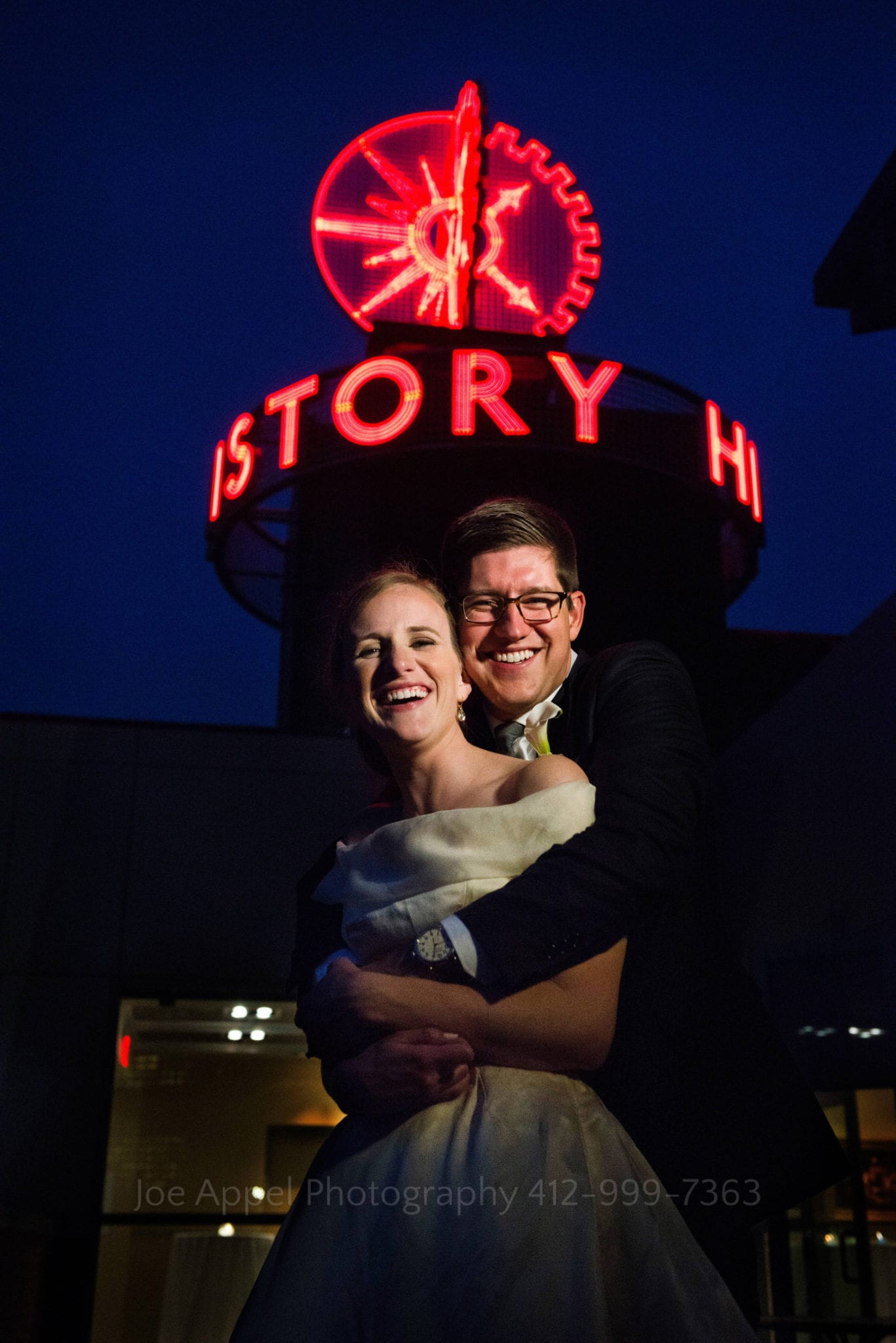 Bride and groom embrace and smile at the camera with the neon Heinz History Center sign above them in the twilight sky.