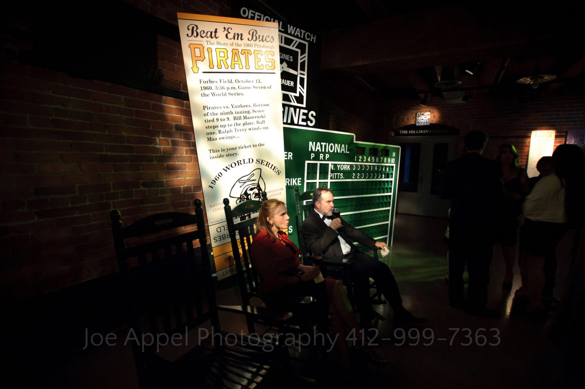 Couple sits in rocking chairs and drinks wine beneath an exhibit on the 1960 world series