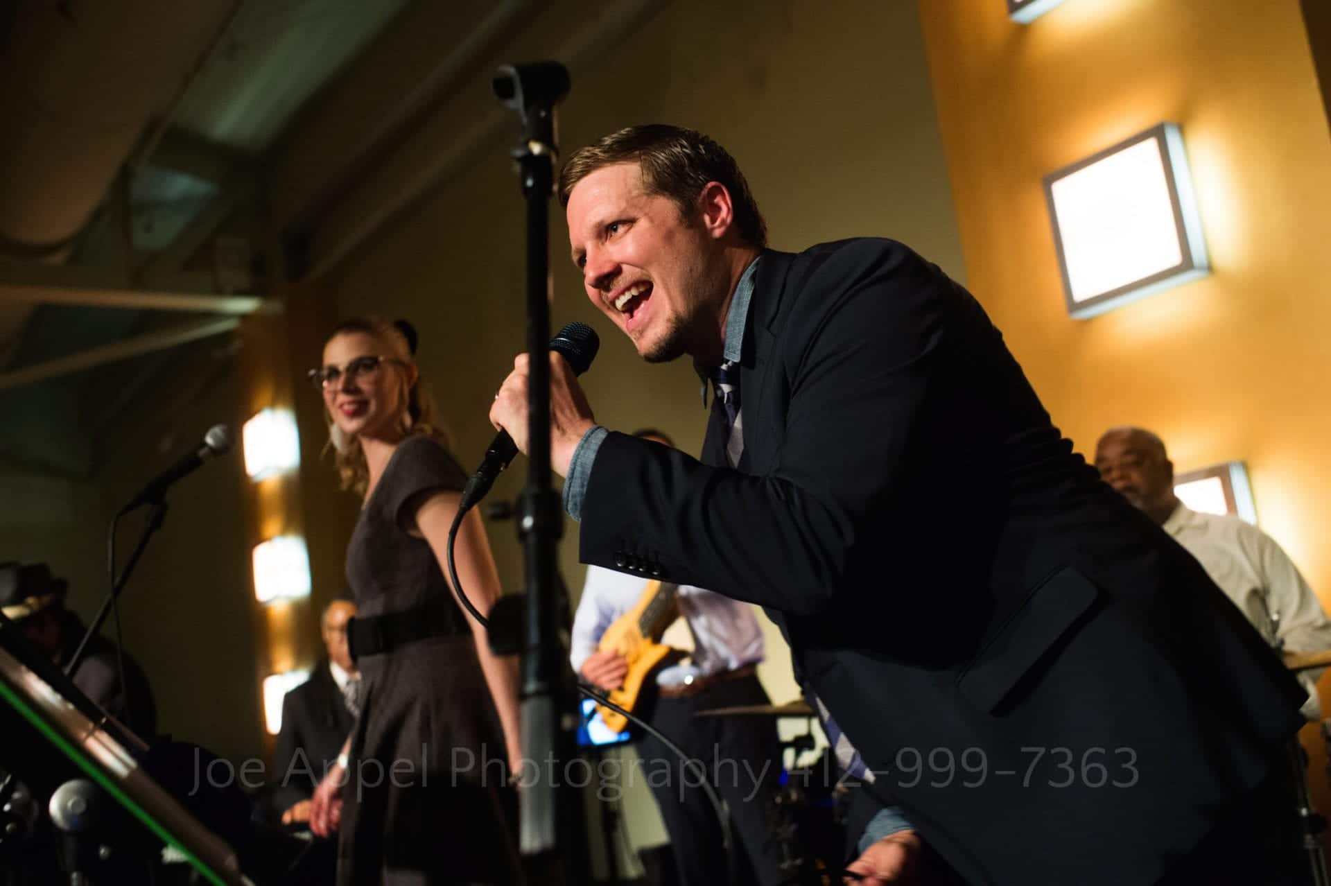 Singer in a band leans forward as he hits a note while a female singer dances behind him.