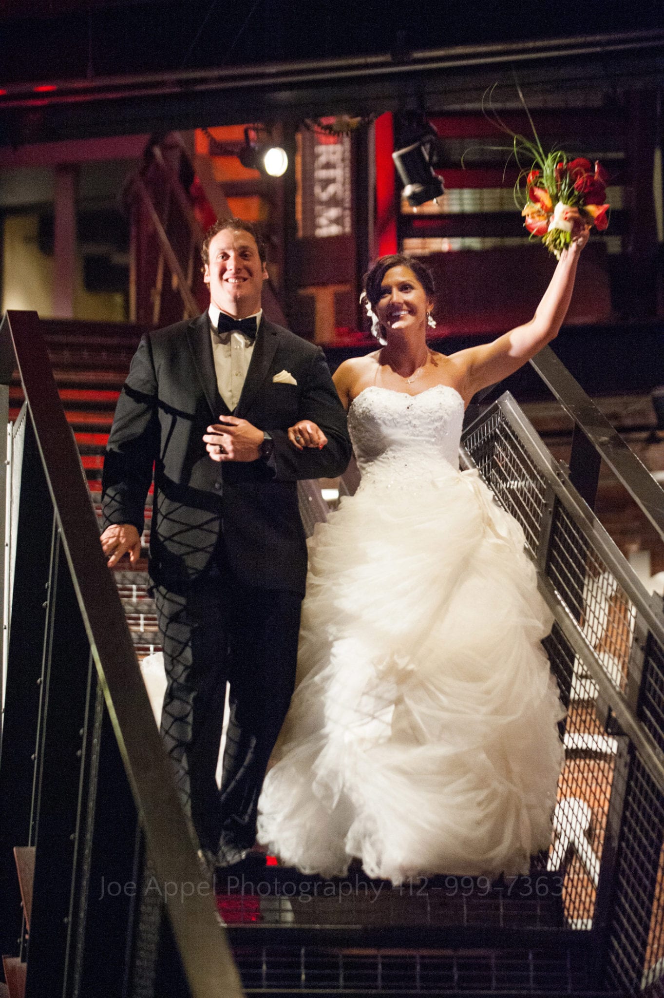 A bride and groom walk down the stairway - the bride in a puffy white dress, raising her bouquet - in the Great Hall at the Heinz History Center