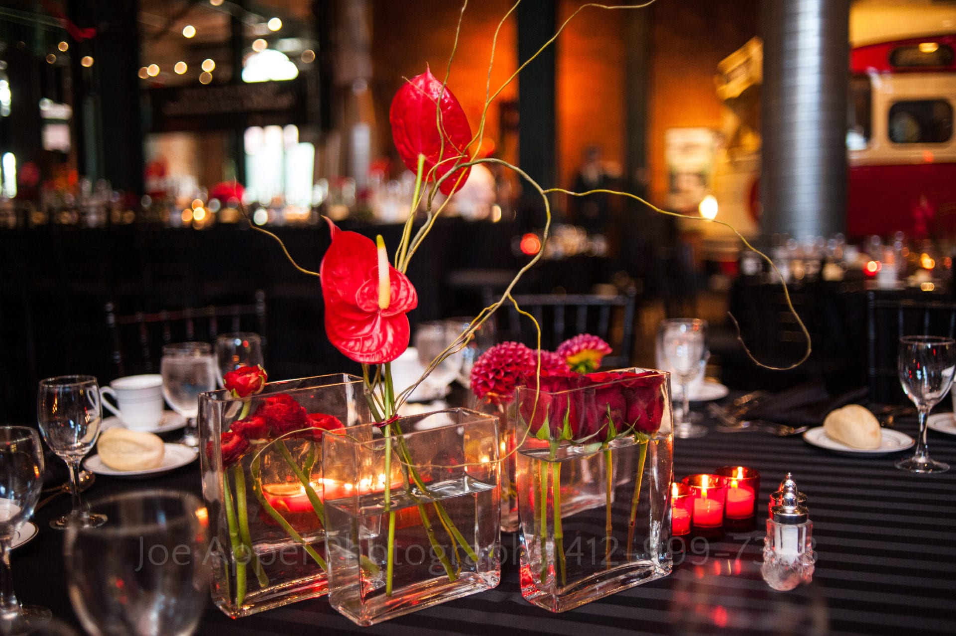 square glass vases contain red roses at the center of a table.