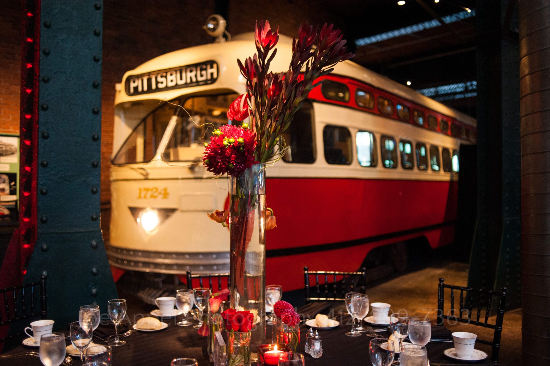 A tall glass vase containing red flowers on a table in front of a Pittsburgh trolley