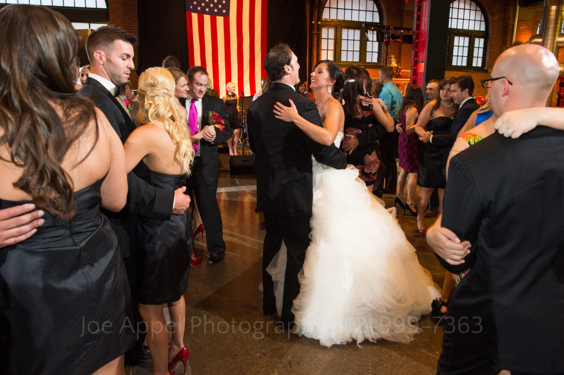 A bride and groom dance in front of a large American flag while other couples fill the dance floor around them.