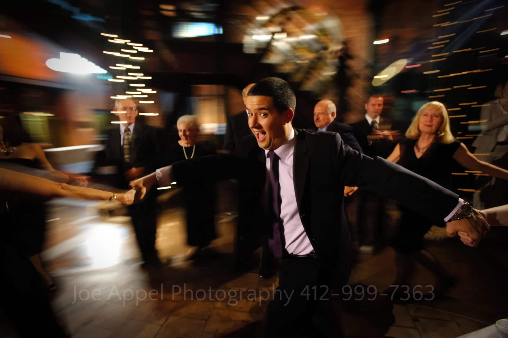 A man yells as he holds hands and dances in a circle with other wedding guests. The background is blurred and shows motion.