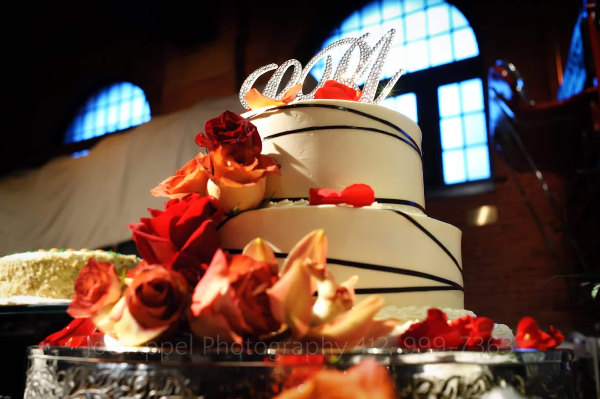 A white cake with red and orange flowers. The crystal initialed topper shimmers in the light. There's a blue window behind the cake.