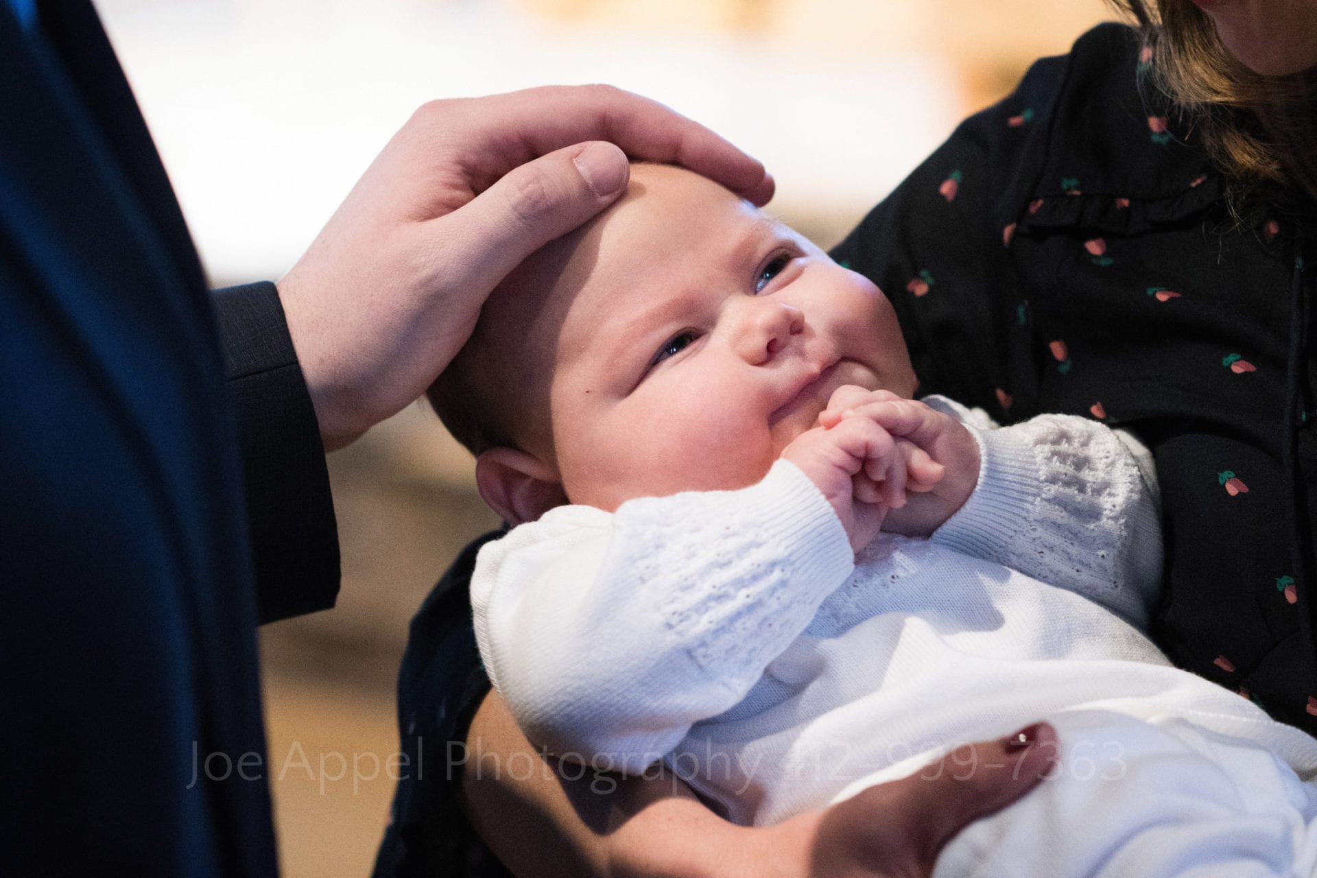 An infant in a white sweater folds his hands together and seems to smile as a man's hand rests on his forehead during a Baptism at Saints John and Paul Parish.